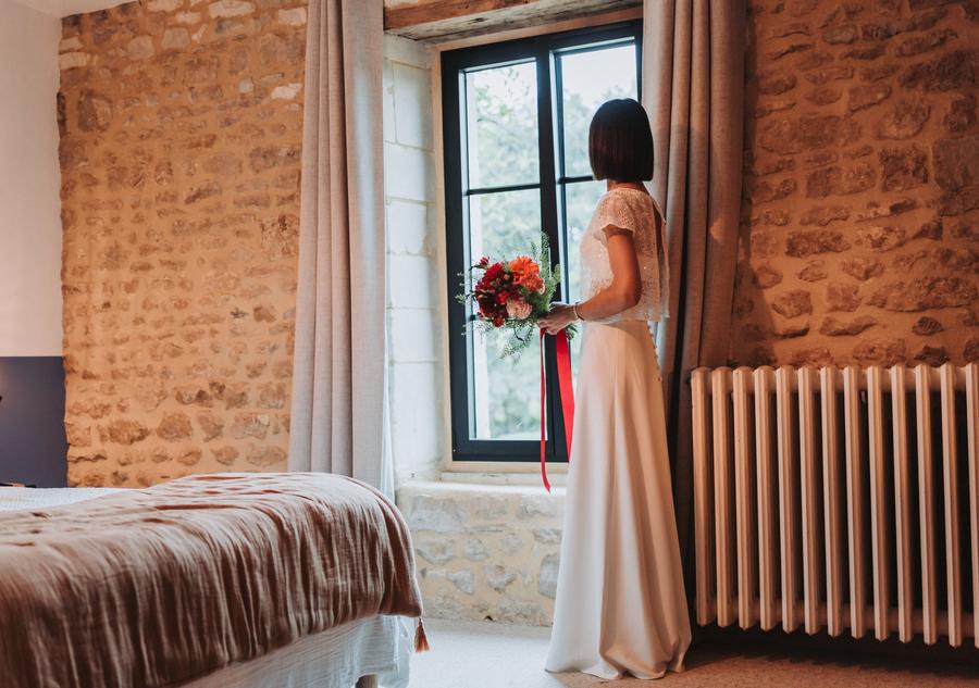 La mariée regarde par la fenêtre, son bouquet rouge à la main